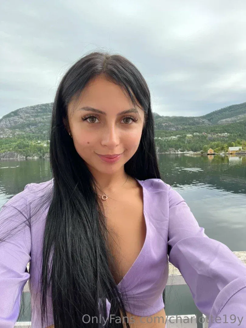 Frau in einem hellvioletten Oberteil, die an einem See mit Hügeln im Hintergrund ein Selfie macht