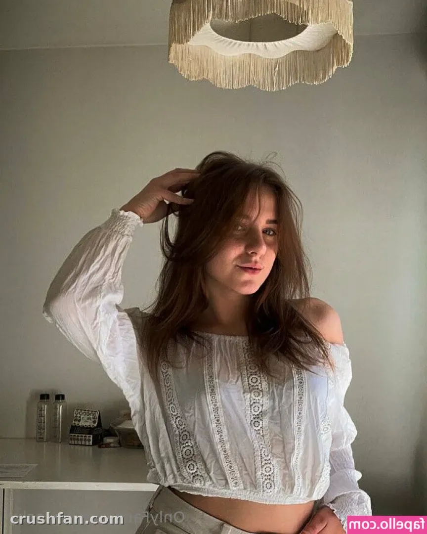 Mulher com blusa branca de renda ombro a ombro ajeitando o cabelo em um ambiente com luz suave