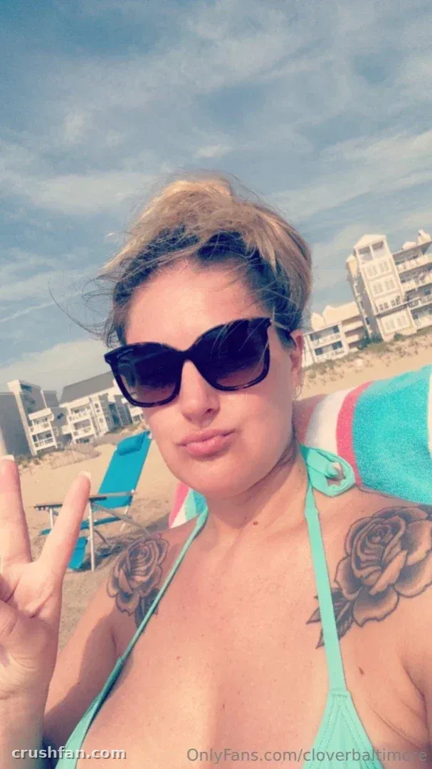 Cloverbaltimore in a teal bikini and sunglasses on a sunny beach, making a peace sign.