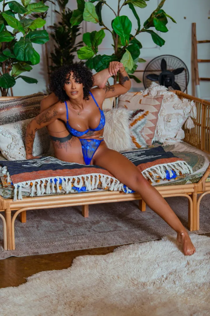 Miss B. Nasty in blue lingerie seated on a rattan daybed surrounded by plants