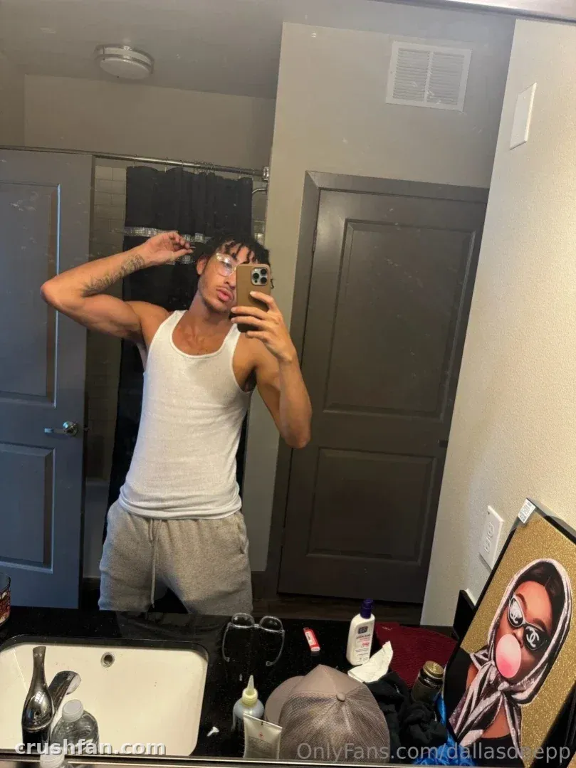 Dallas en débardeur blanc et pantalon de jogging gris prenant un selfie dans le miroir d'une salle de bain.