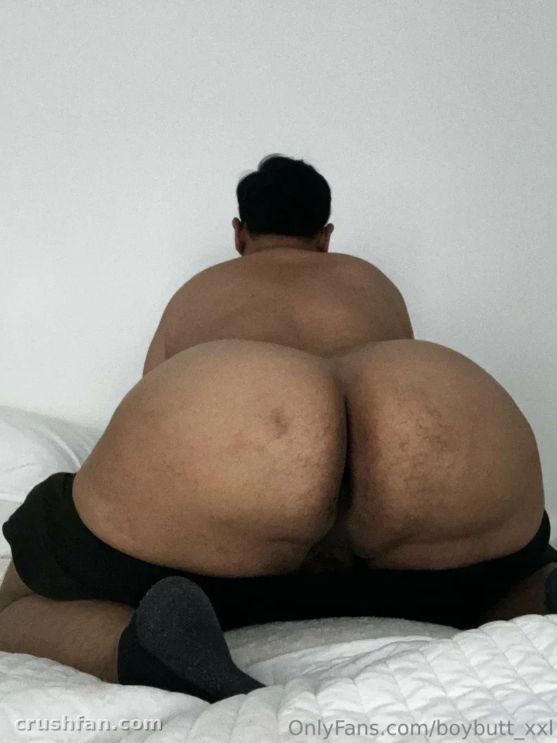 BoyButtXXL kneeling on a white bed in black underwear against a plain white wall