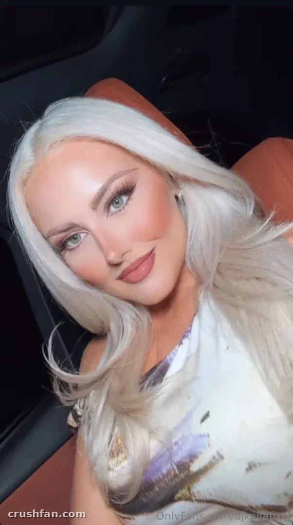 Kaila Troy com longo cabelo loiro platinado em uma blusa branca com estampa dentro de um carro