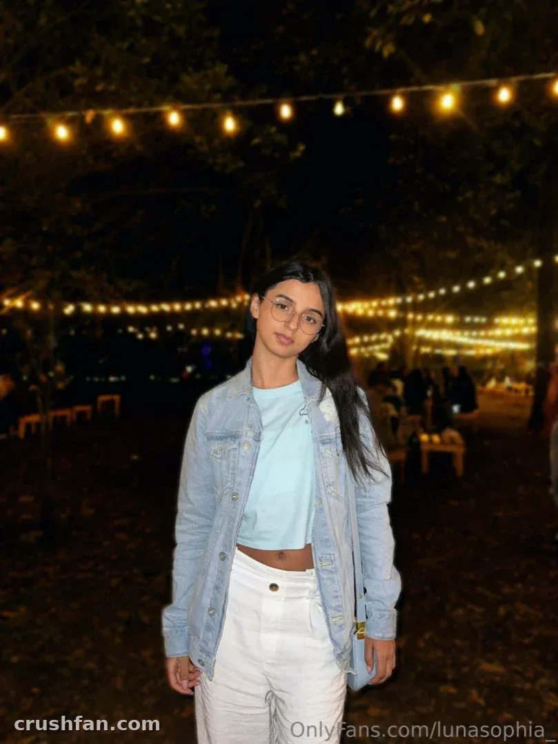 Luna Sophia in a denim jacket and white trousers under warm string lights at night