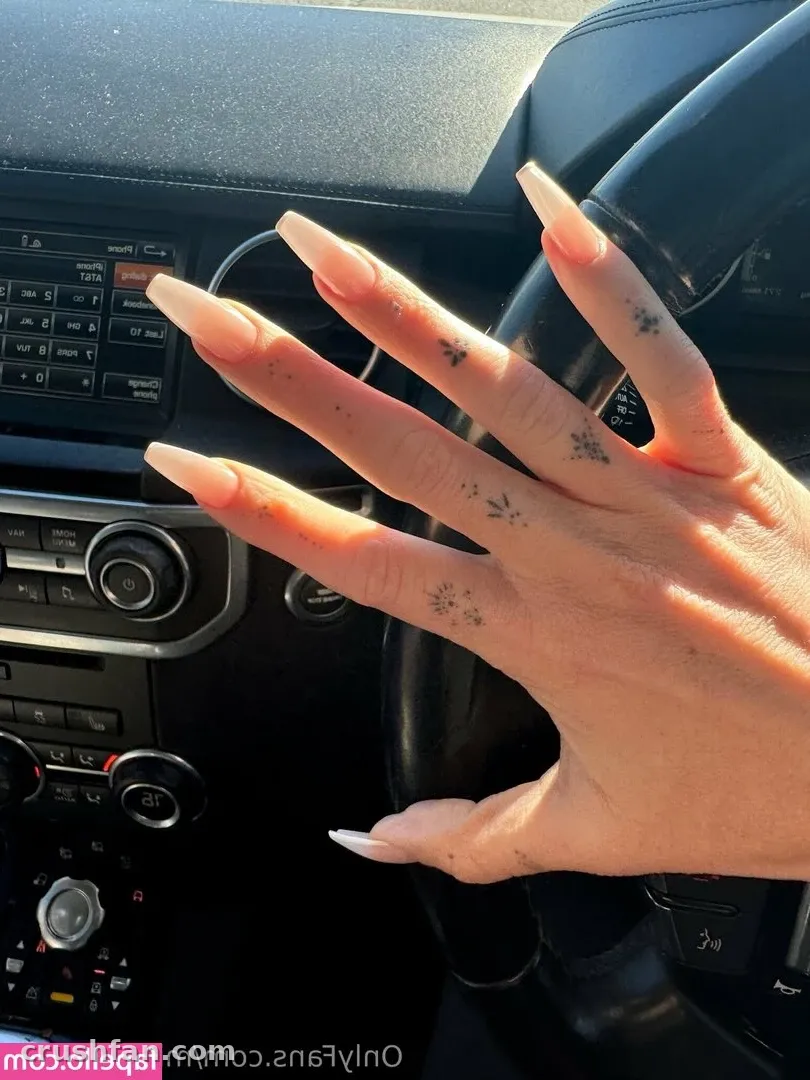 Hand with long pink nails and small tattoos resting near a steering wheel in a car