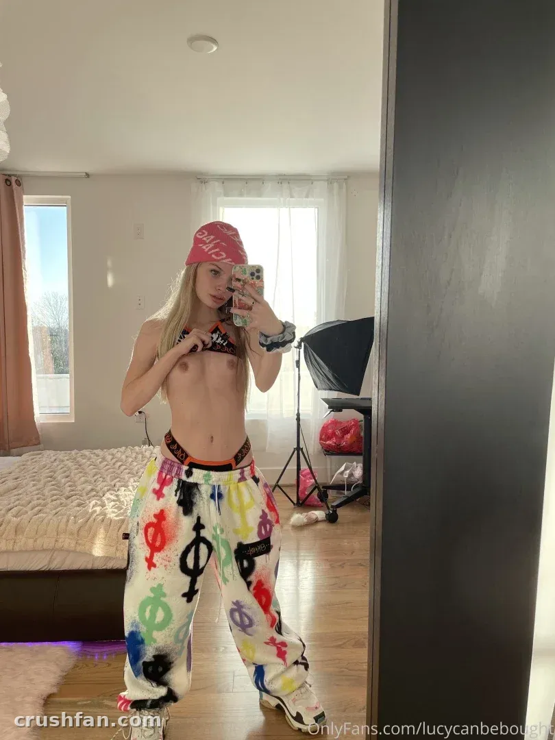 Lucy con gorra rosa y pantalones de estampados coloridos tomándose una selfie frente al espejo en un dormitorio luminoso
