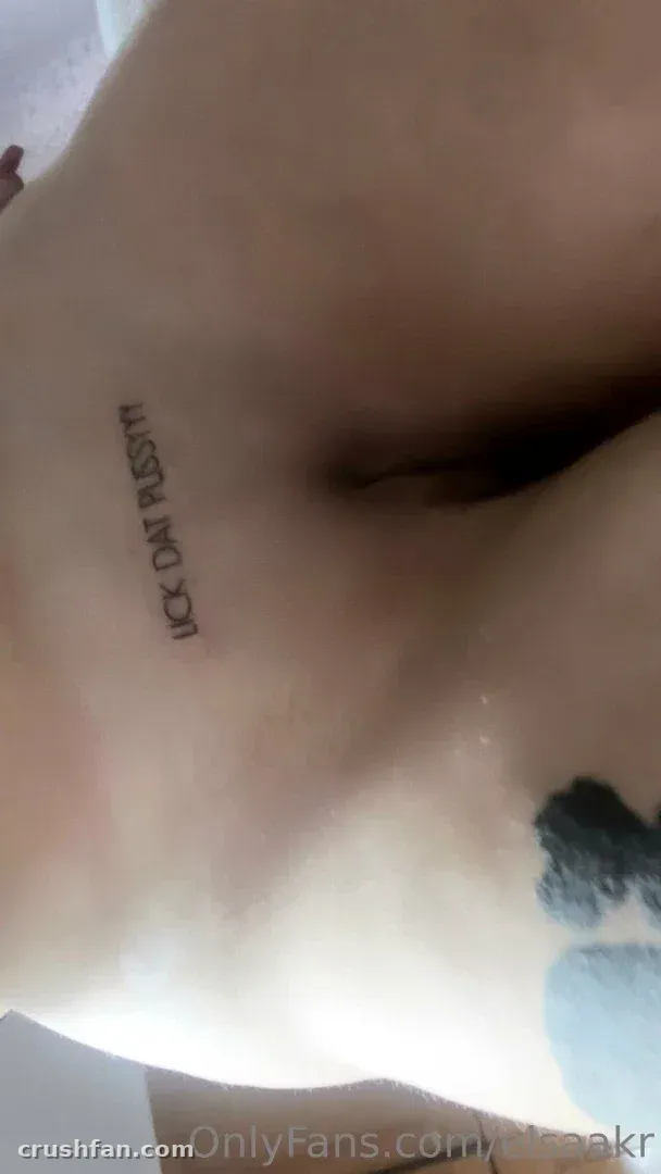 Close-up da clavícula de Elisabeth com uma pequena tatuagem, suavemente iluminada contra um fundo pálido