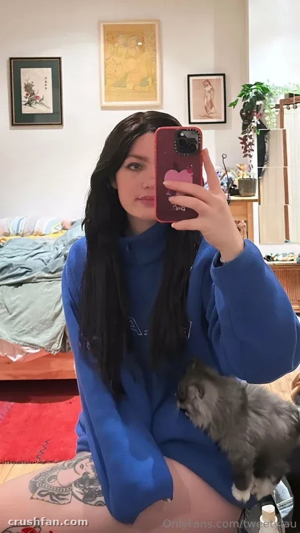 Laura in a blue hoodie taking a mirror selfie with a grey cat beside her in a bedroom.