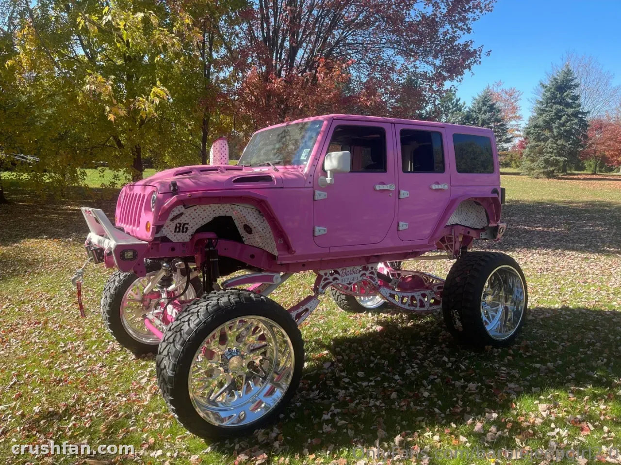 Jeep elevado rosa com rodas cromadas estacionado em um gramado coberto de folhas sob árvores de outono