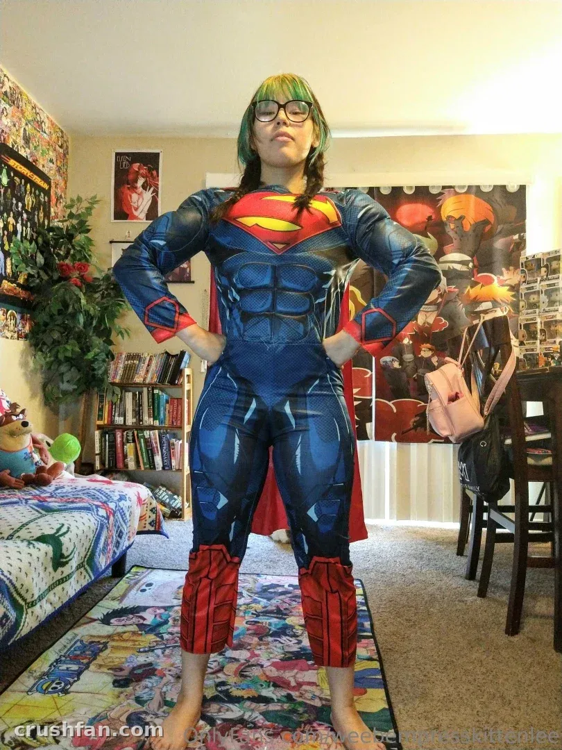 EmpressKoneko in a blue superhero costume standing in a bedroom with a bookshelf and poster.