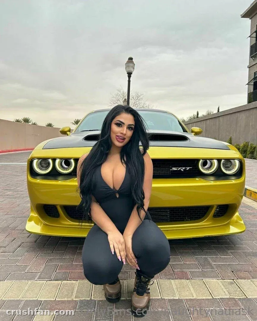 Nadia crouching in a black outfit in front of a yellow sports car on brick pavement