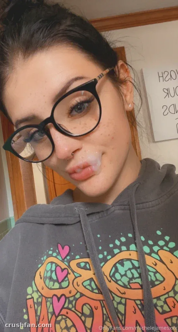 Michele James com um moletom cinza estampado e óculos pretos tirando uma selfie em ambiente interno