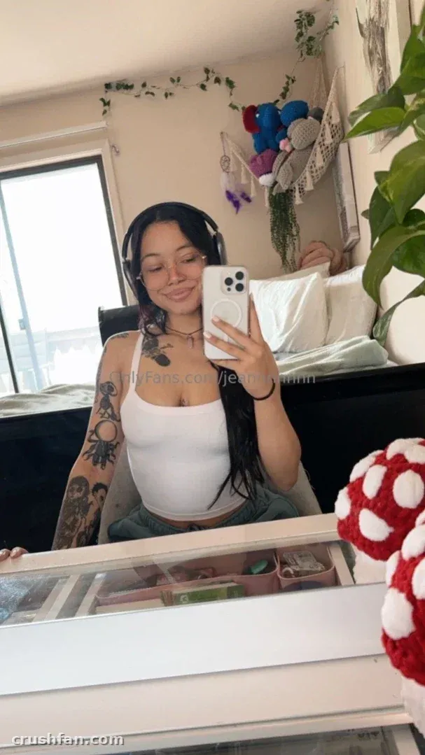 Jean in einem weißen Top und mit Kopfhörern, wie sie in einem Schlafzimmer ein Spiegel-Selfie macht.