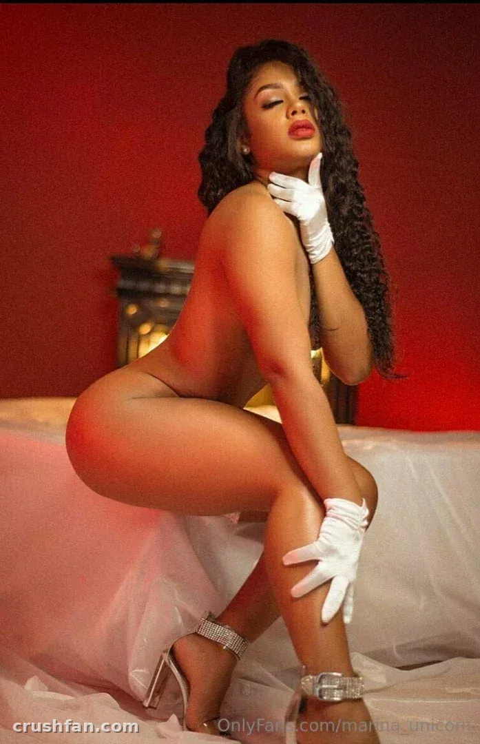 Woman with dark curled hair kneeling on white sheets against a red wall, wearing white gloves and heels