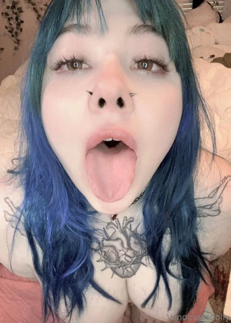 Venomous Dolly with blue hair sticking out her tongue, showing chest tattoos in a bedroom.