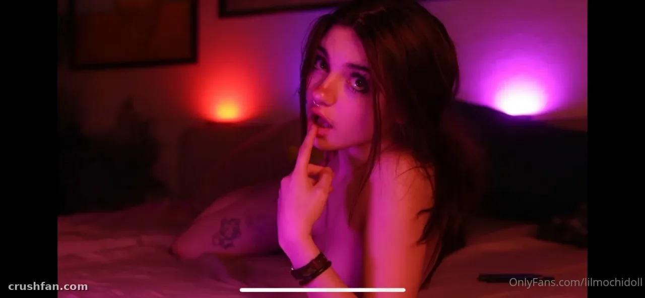 Lil Doll resting on a bed in a room lit by magenta and violet light
