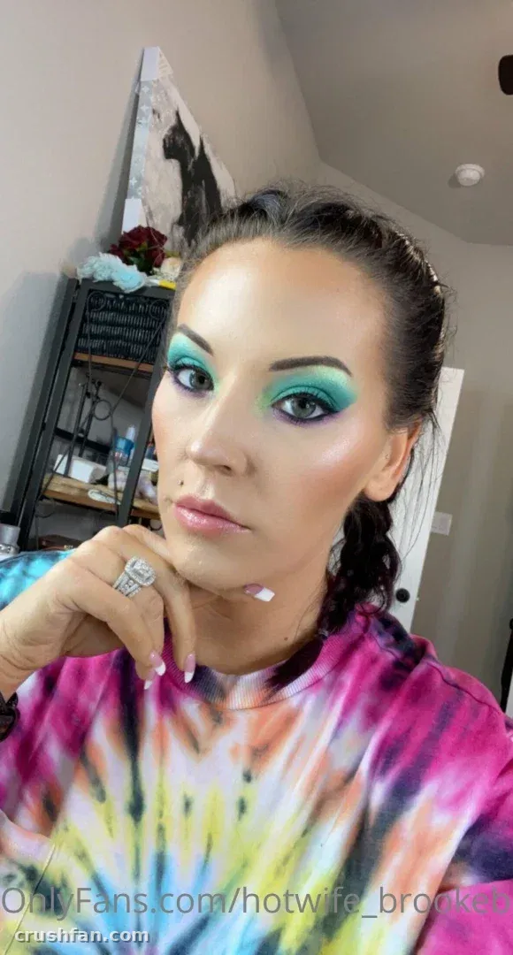 Creator in einem farbenfrohen Batik-Oberteil mit auffälligem türkisfarbenem Augen-Make-up, drinnen.