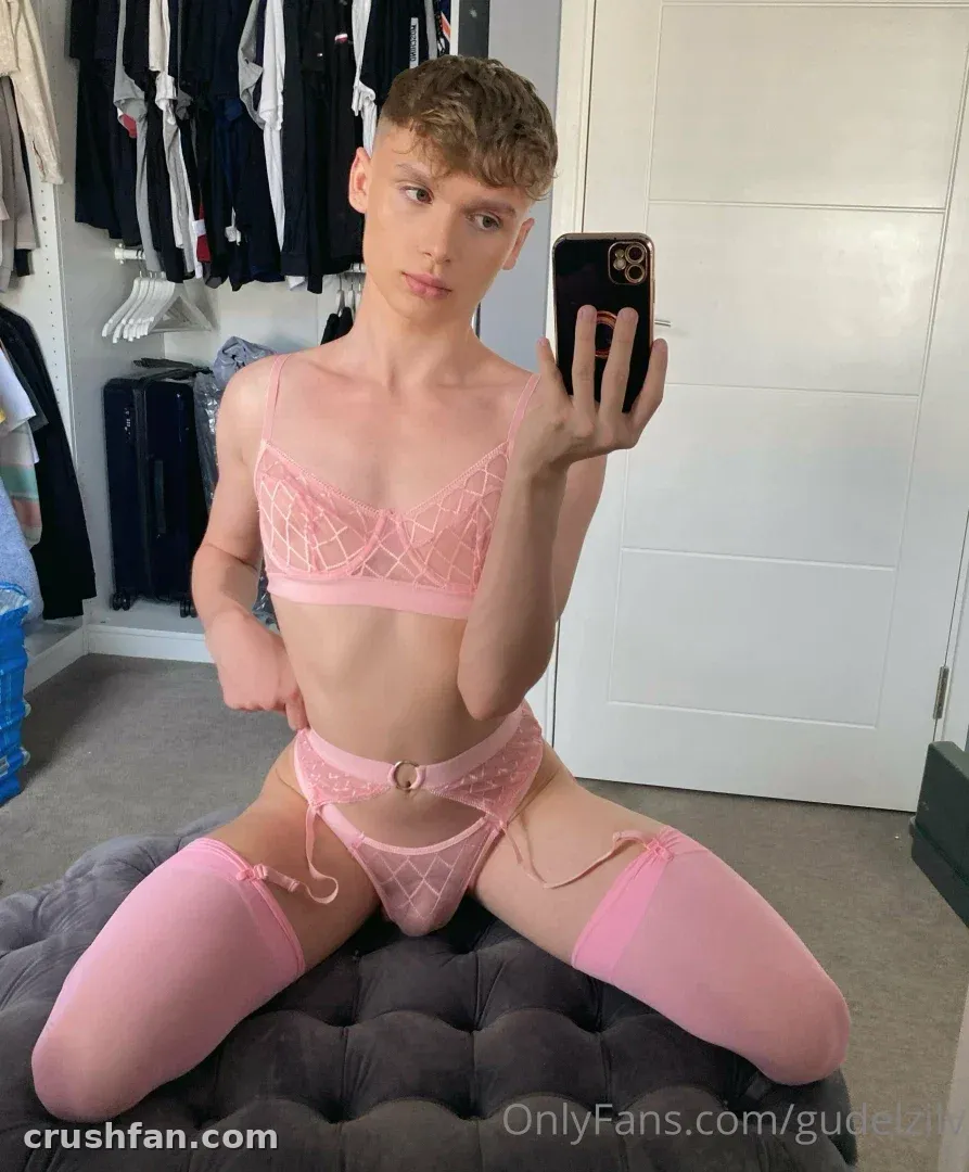 Criador em lingerie de renda rosa sentado em um pufe cinza tirando uma selfie no espelho em uma sala com um armário.
