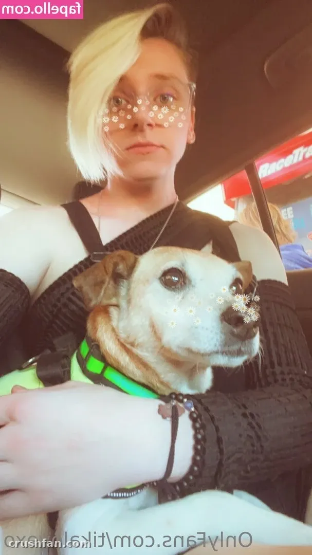 Tika in a black knit top holding a small dog wearing a green harness in a casual selfie
