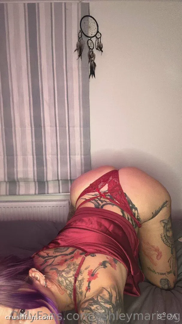 AshleyMarie в красном белье на кровати с видимыми татуировками, ловец снов на стене позади