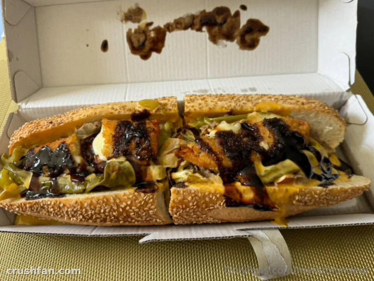 Um sanduíche sub com molho em uma caixa de takeaway aberta sobre uma superfície verde-oliva.