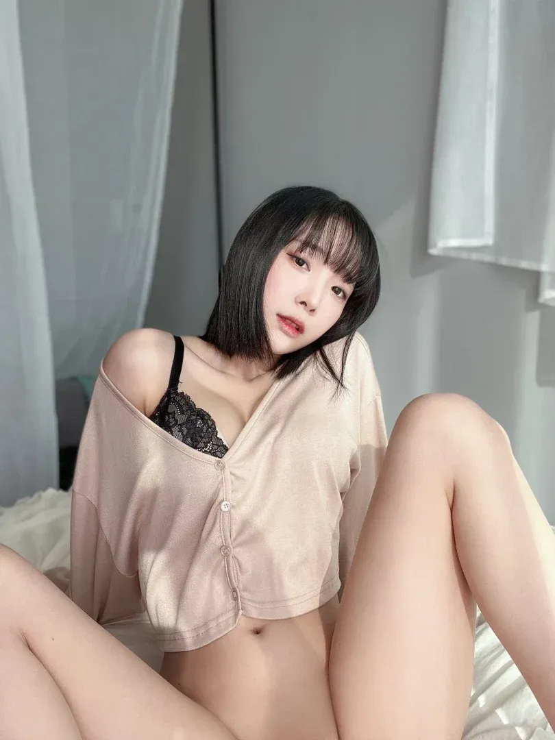 inkyung97 in a beige shirt and black lace top sitting on a white bed