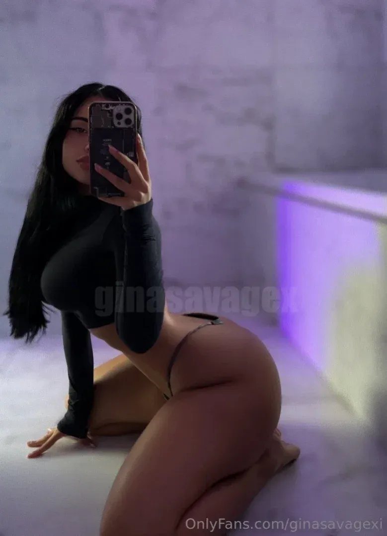 Ginasavagex agenouillée sur un sol carrelé prenant un selfie dans le miroir avec un haut noir et un bas de bikini sous une lumière violette.