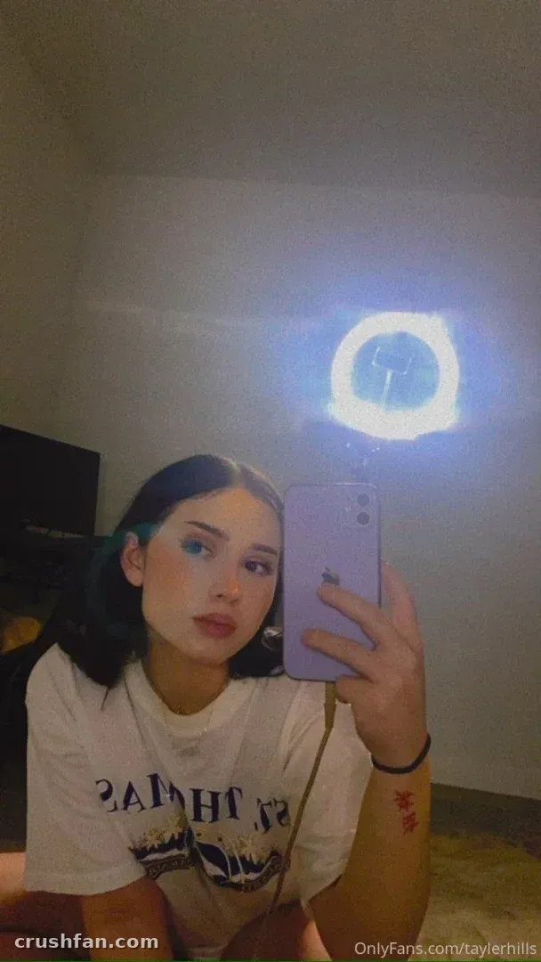 Tayler hills taking a mirror selfie in a white graphic tee with a ring light behind her