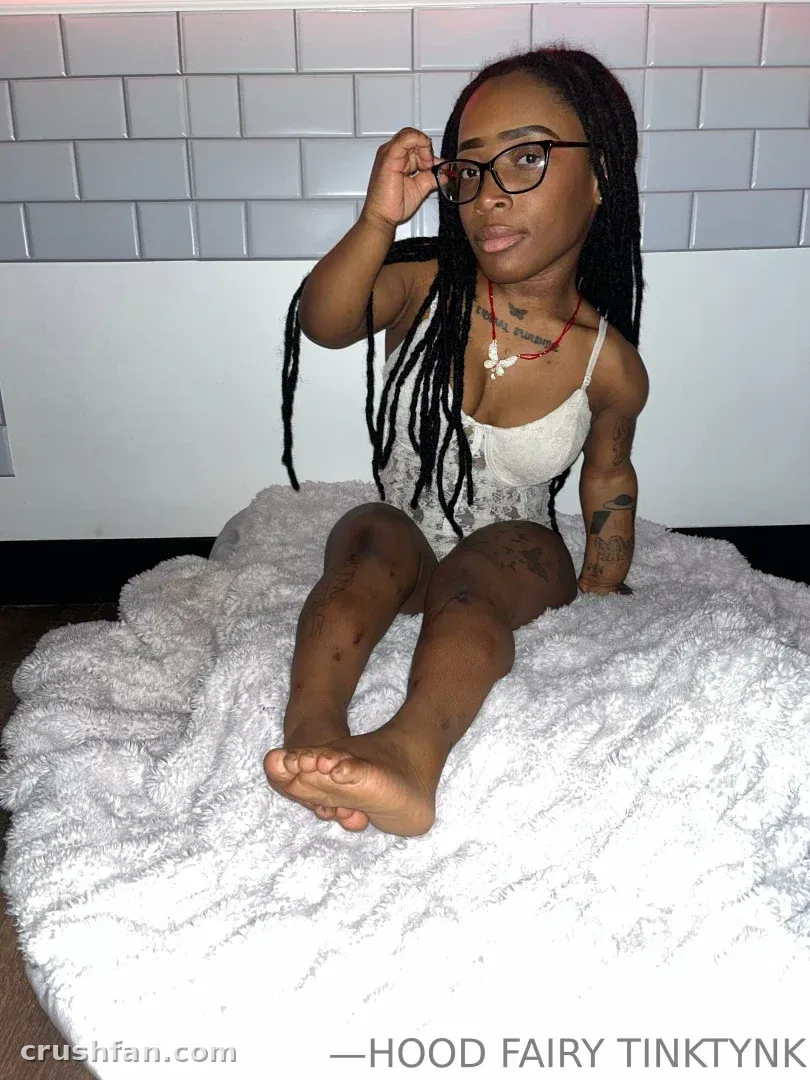 Tinky Winky sitting on a white blanket wearing glasses and a lace top indoors