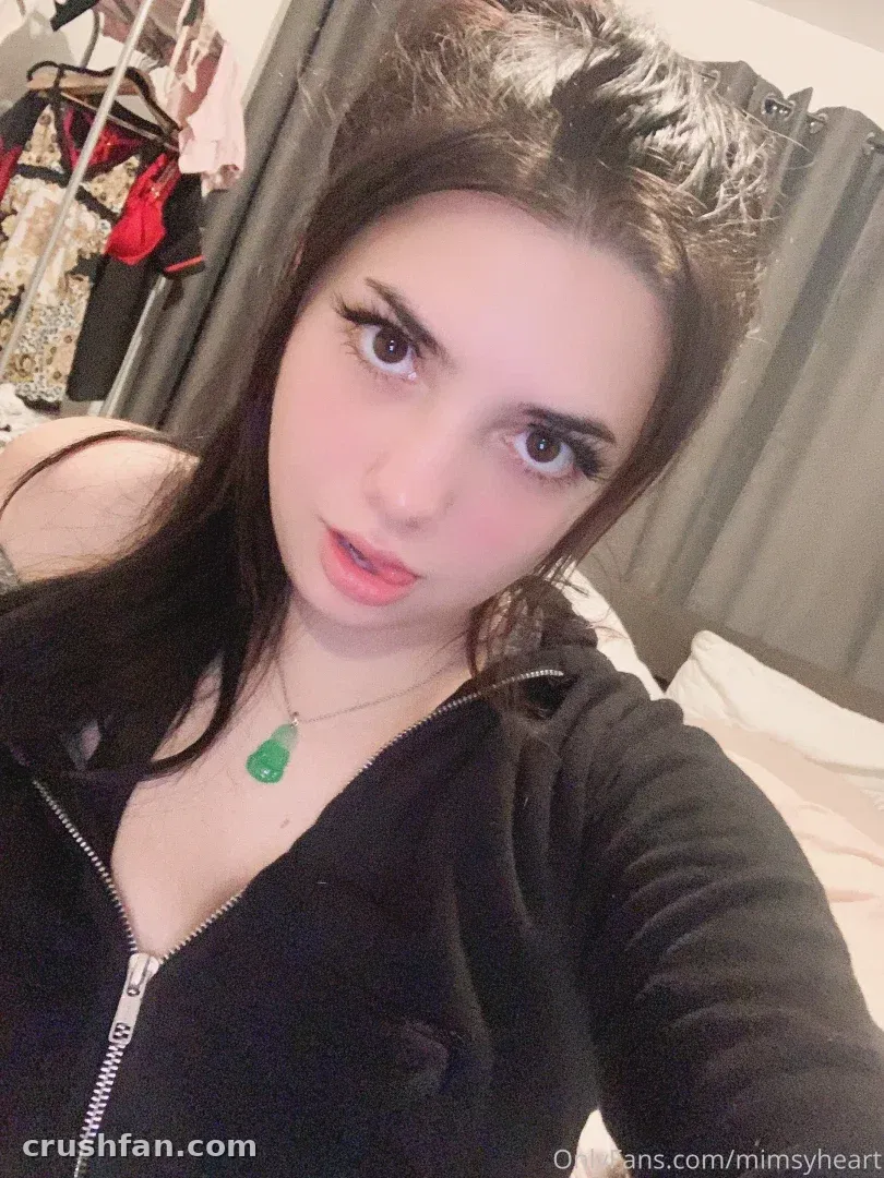 Amber em um top preto com zíper e um pingente de jade, tirando uma selfie em ambiente interno.