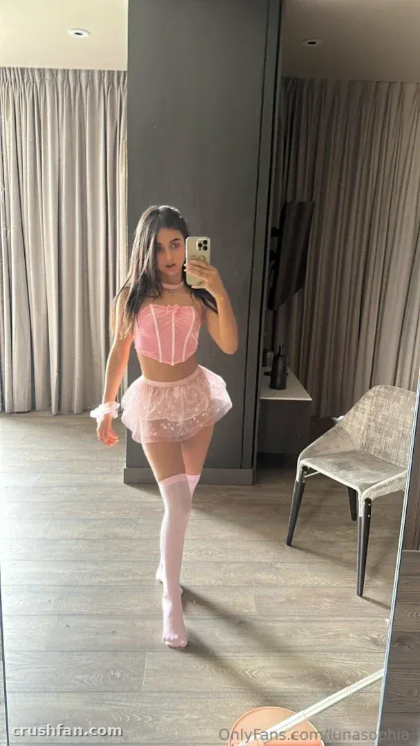Luna Sophia tirando uma selfie no espelho com um corset e saia rosa e meias brancas.