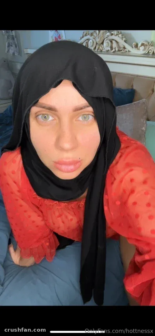 Criadora com hijab preto e blusa vermelha de bolinhas posando em ambiente interno para um retrato selfie próximo.