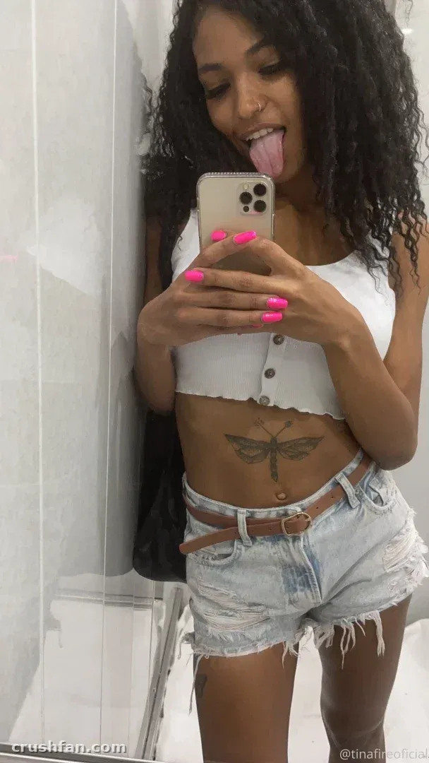 Tina Fire taking a mirror selfie in a white crop top and denim shorts.