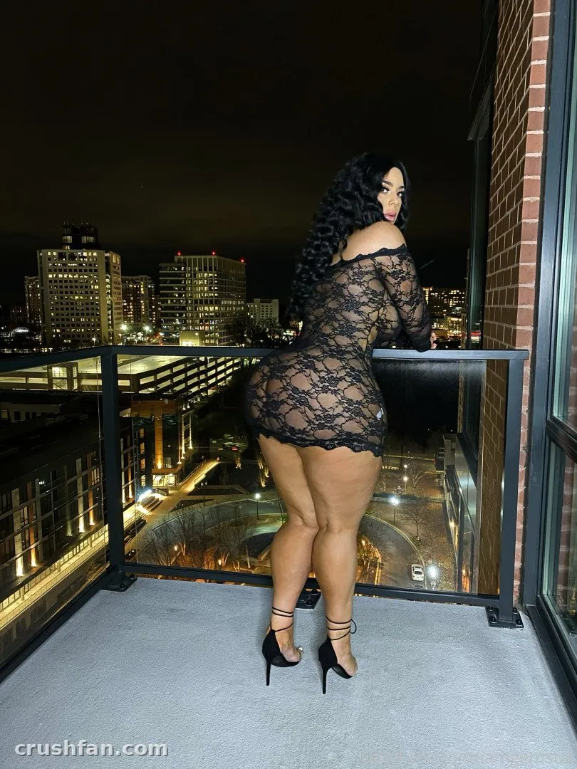 Gemstar com um vestido de renda preto e saltos em uma varanda com vista para uma cidade à noite.