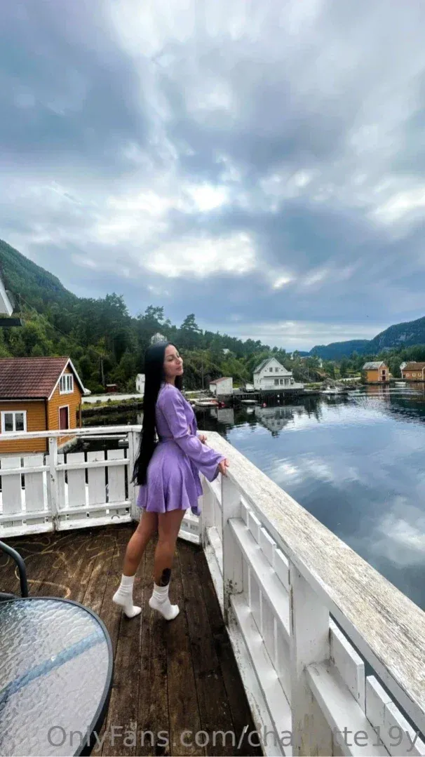 Mulher de vestido roxo em pé ao lado de um corrimão branco em um deck de madeira com vista para a água e colinas.