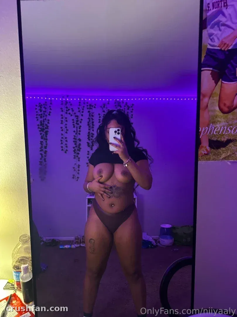 Creator in a black two piece taking a mirror selfie in a purple lit room with hanging vines.