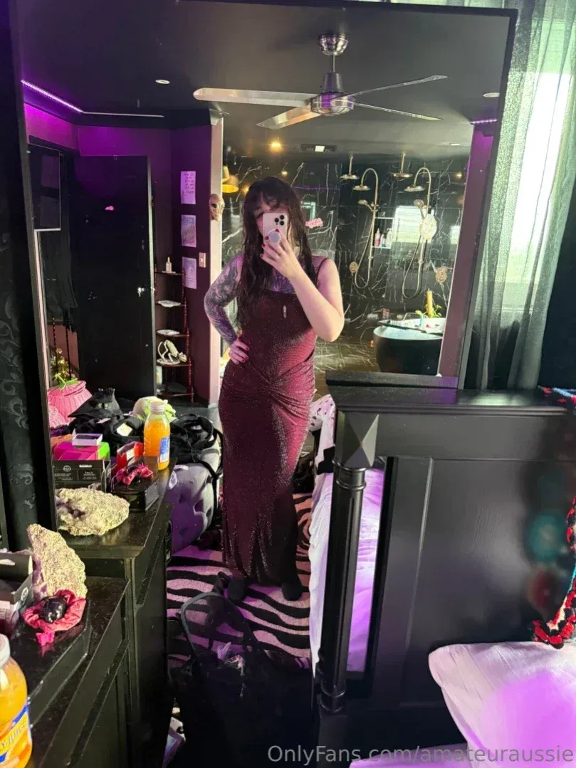 Aus Sarah com um vestido longo na cor vinho tirando uma selfie no espelho em um quarto com pouca luz