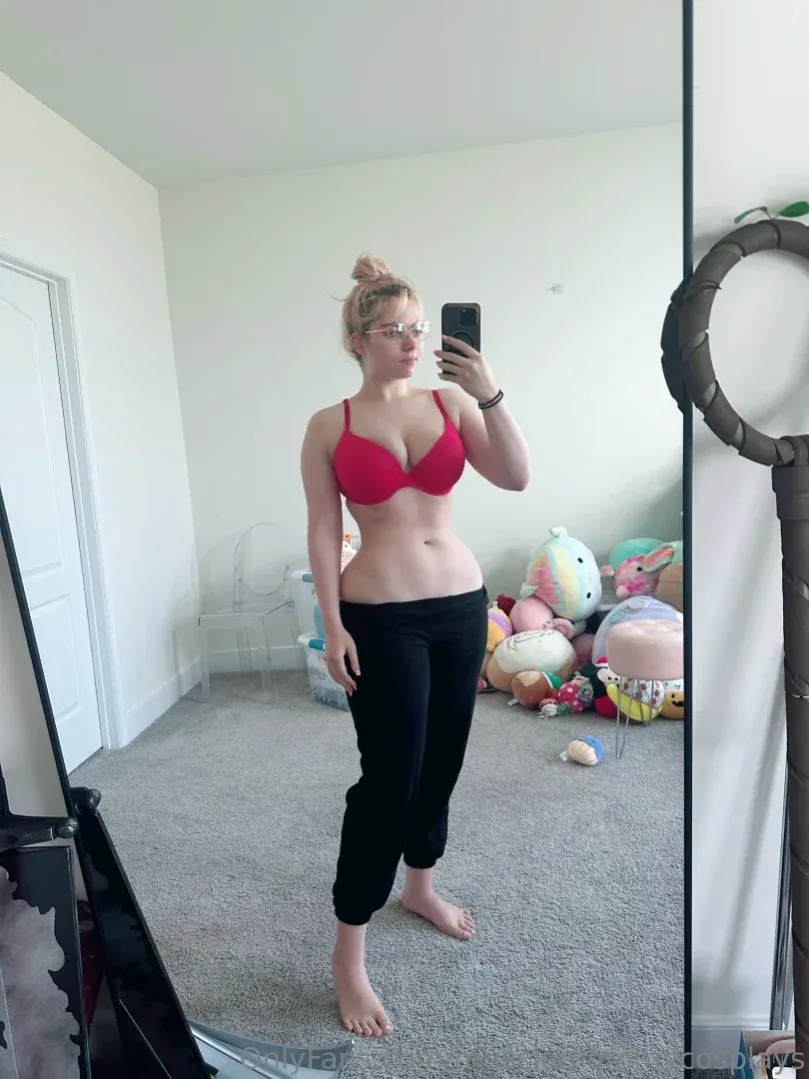 Linnea tira uma selfie no espelho com um sutiã vermelho e calça preta cropped em um quarto claro.