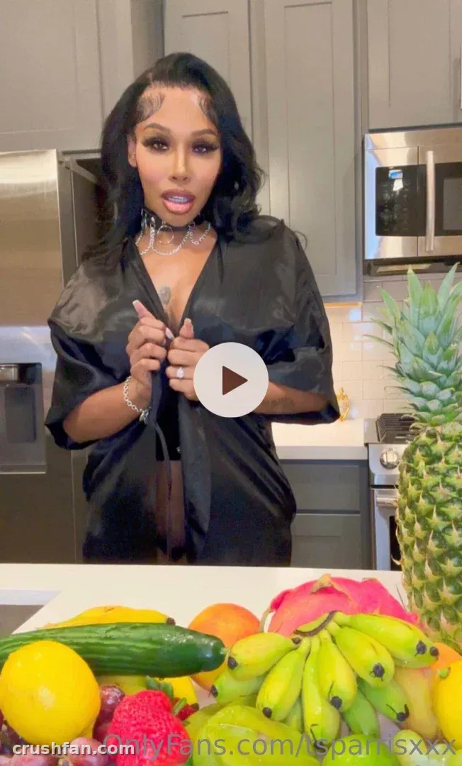 Creator in a black satin robe standing behind a counter of fresh fruit in a modern kitchen.