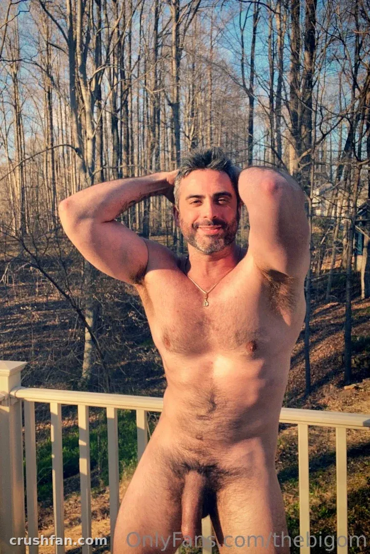 Hombre desnudo con los brazos levantados detrás de la cabeza de pie en una plataforma de madera ante árboles desnudos.