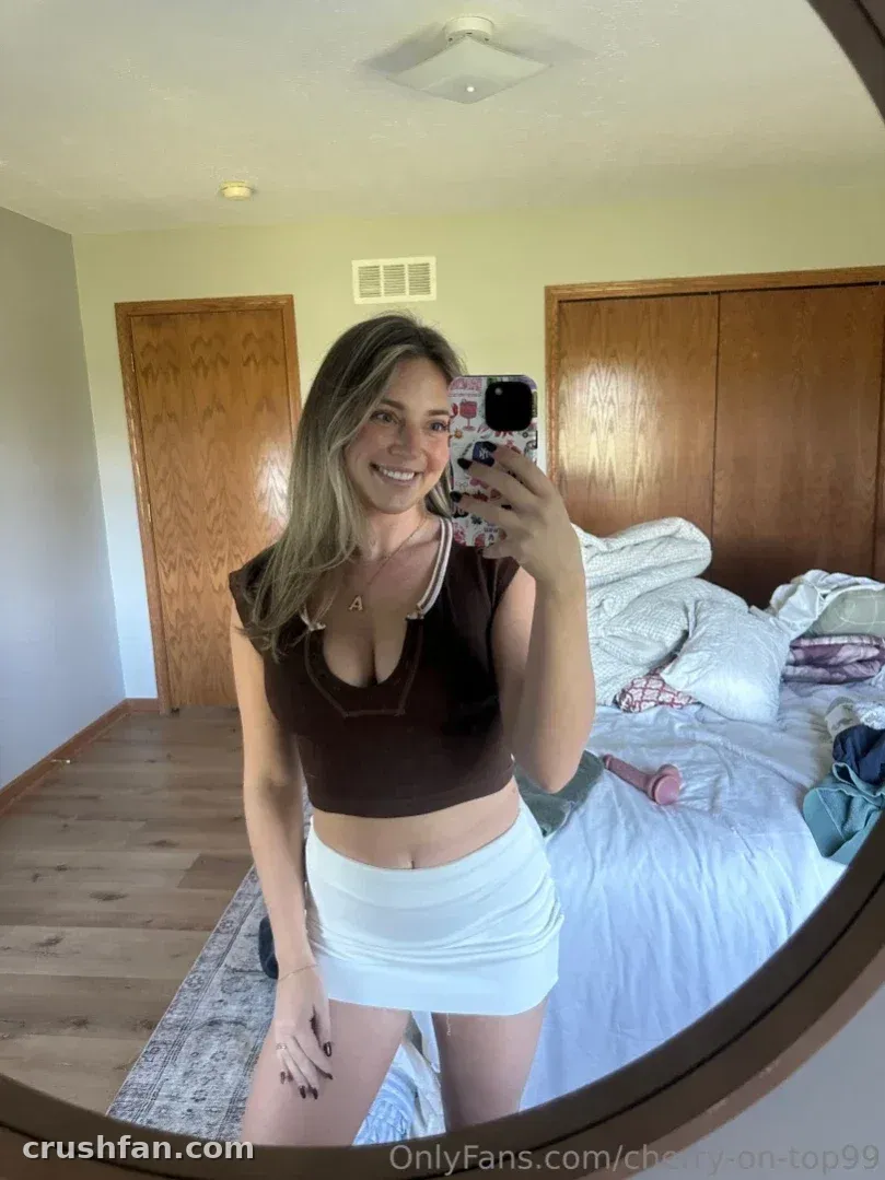 Cherry-on-top taking a mirror selfie in a brown crop top and white skirt inside a bedroom.