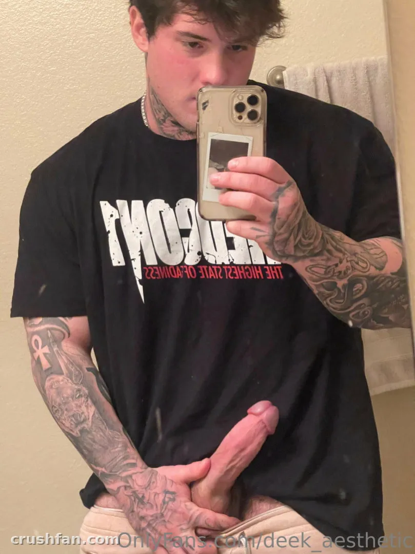 DEEK AESTHETIC macht ein Spiegel-Selfie in einem schwarzen Grafik-T-Shirt in einem Badezimmer.