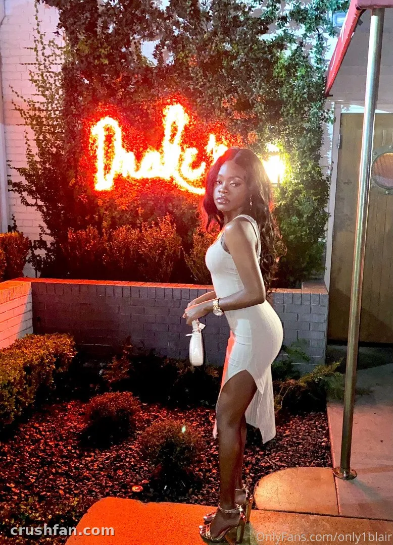 Blair in a white dress standing beside a glowing neon sign and green foliage at dusk