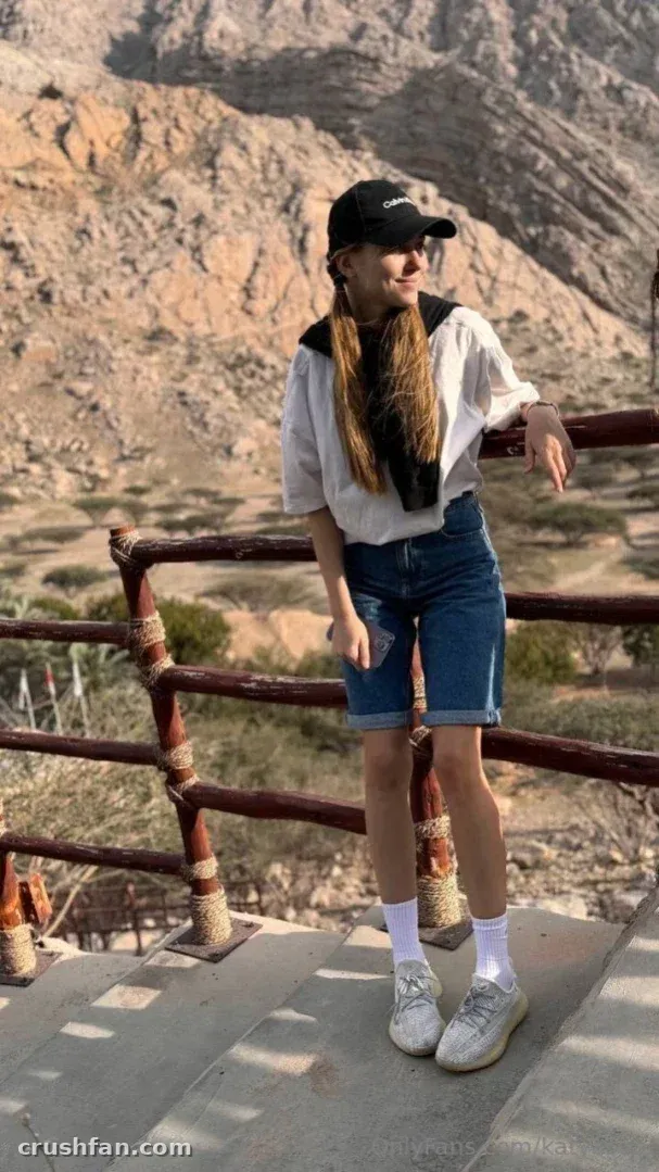 Katya Sun com uma blusa branca e shorts jeans em pé ao lado de uma grade de madeira em uma trilha de montanha.