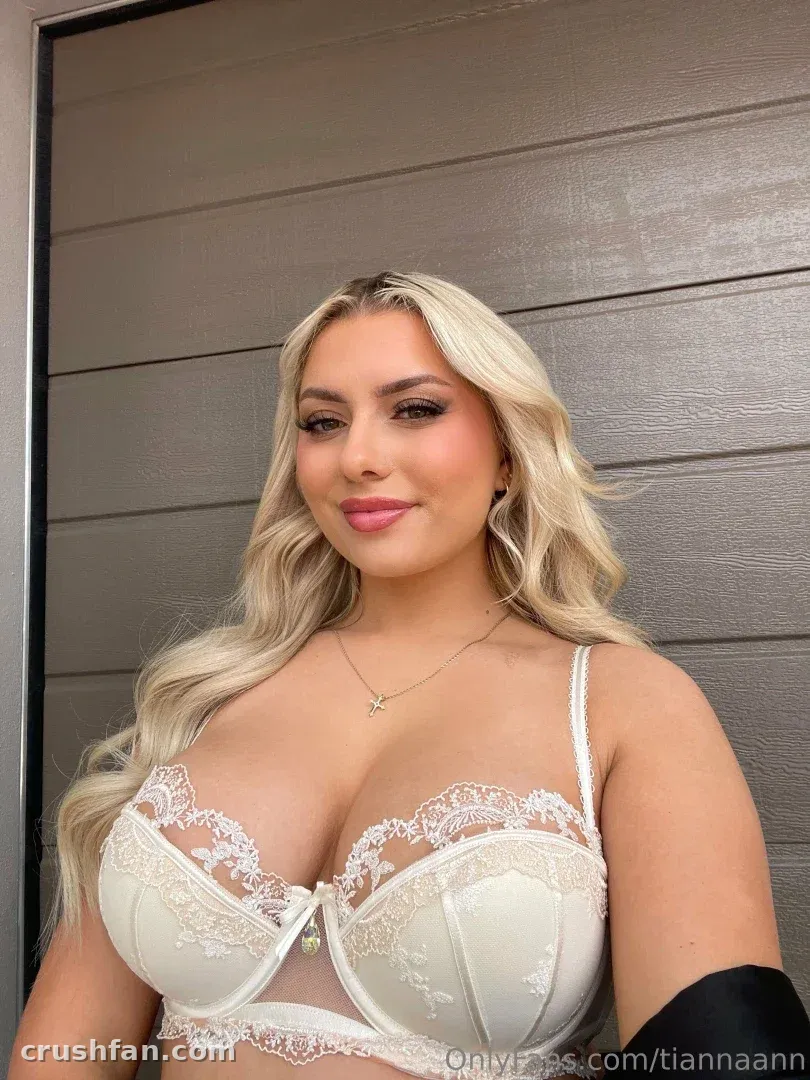 Tianna com um conjunto de lingerie de renda branca, sorrindo para a câmera diante de uma parede cinza com painéis
