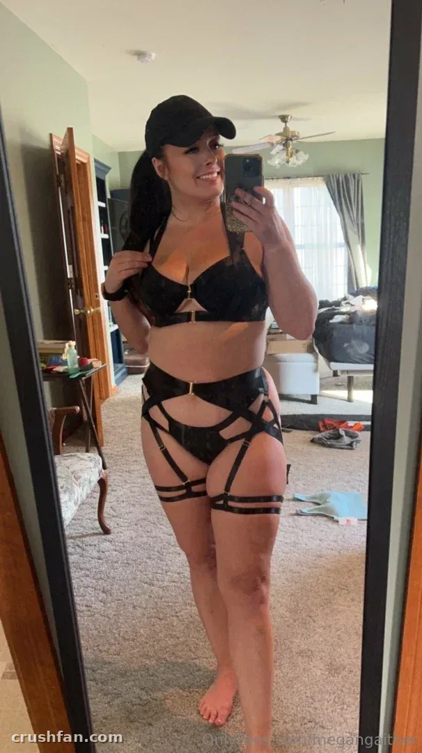 Megan Gaither de lingerie preta e boné tirando uma selfie no espelho em um quarto com carpete