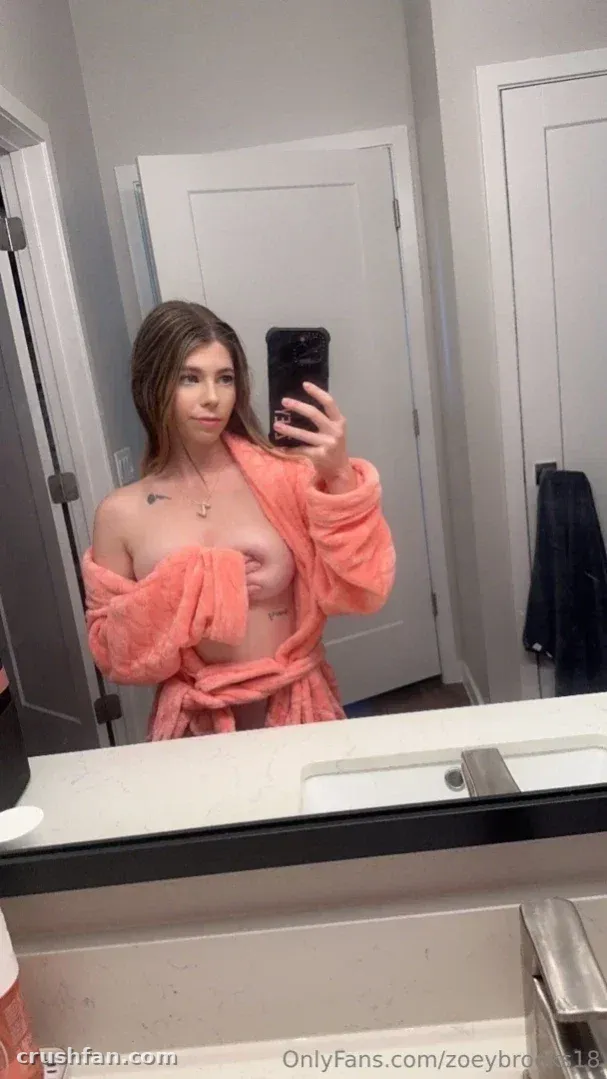 Zoey Brooks tomándose una selfie frente al espejo con una bata coral en un baño