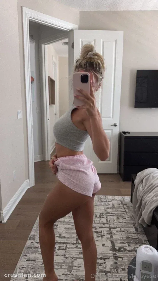 Kyla prend un selfie dans le miroir en crop top gris et short rose dans une pièce lumineuse
