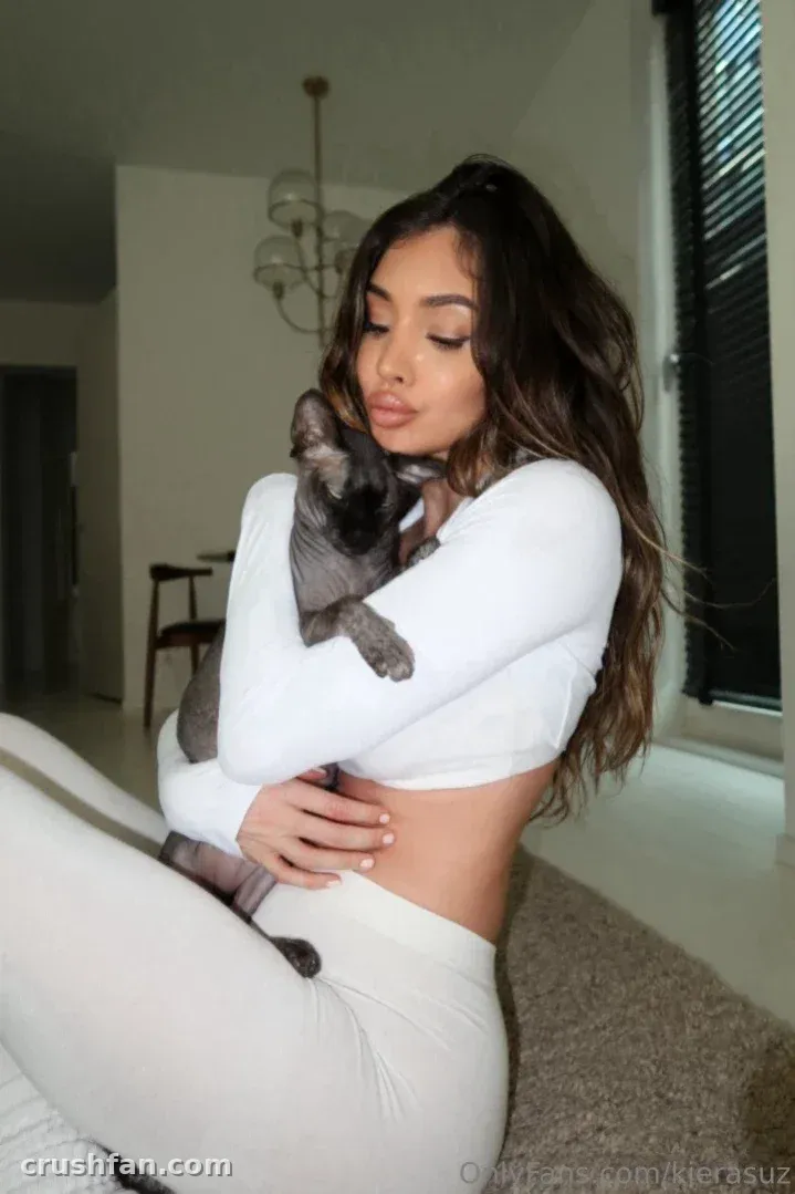 Kiera com um top cropped branco e leggings segurando um gato cinza dentro de casa.