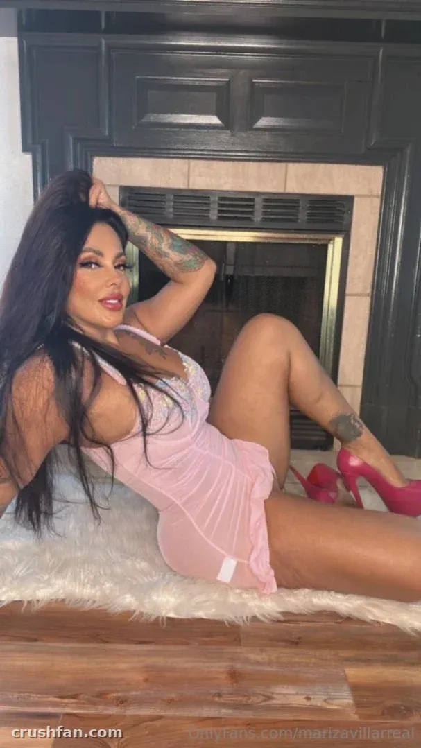 Mariza Villarreal dans un body en dentelle rose et des talons roses allongée sur un tapis en fourrure blanche près d'une cheminée.